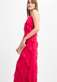 Langes rosa Kleid mit geschichteten Rüschen, Spaghetti-Trägern und fließender Silhouette. Das Model hält eine goldene Clutch, die die Eleganz des Kleides unterstreicht.
