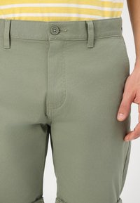 Pantaloni verzi din bumbac cu o textură netedă, având închidere cu buton, bucle pentru curea și detalii de cusătură discrete. Croială adaptată în jurul taliei.