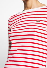 Maglia a maniche lunghe a coste con righe orizzontali rosse e bianche, caratterizzata da un piccolo logo di coccodrillo ricamato in verde sul petto.