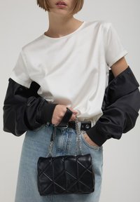 Borsa a tracolla nera trapuntata con tracolla a catena, abbinata a una maglietta bianca in satin e maniche nere in satin, indossata con jeans in denim blu.
