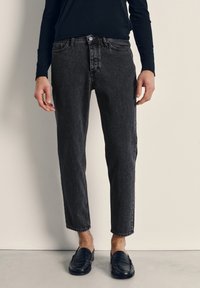 Moški oblečen v črne ozke kavbojke in črne čevlje brez vezalk stoji ob beli steni in sivi tla.