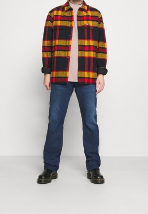Chemise en flanelle à carreaux rouge, jaune et noir, avec deux poches poitrine ; associée à un jean bleu et des bottes noires. Texture de tissu lisse.