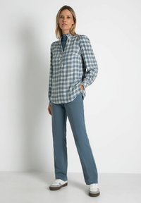 Karierte Bluse in Blau und Weiß, lange Ärmel, Knopfverschluss vorne; kombiniert mit geraden türkisfarbenen Hosen und weißen Sneakers mit braunen Sohlen.
