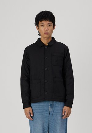 Veste noire à boutons avec un col, deux poches avant et un tissu texturé. Portée avec un jean bleu clair.