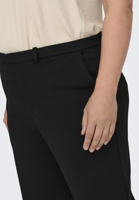 Svarta byxor i ett mjukt, lätt material. Har sidofickor, en platt midja och en skräddarsydd passform. Handen vilar vid sidan.