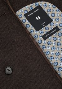 Wnętrze brązowej wełnianej kurtki z jasnobeżową podszewką z niebieskimi wzorami geometrycznymi; zawiera metki z informacjami o marce i materiale.