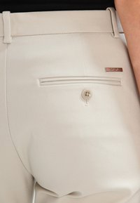 Beige skräddarsydda byxor med en bakficka med horisontell dragkedja och en knäppning, samt en metallisk logotypdekoration.