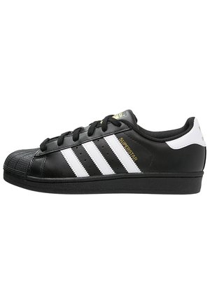 Baskets Adidas Superstar noires avec des bandes blanches, des lacets noirs, un bout en caoutchouc et l’inscription « SUPERSTAR » en doré sur le côté.
