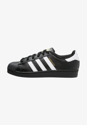 Scarpa da ginnastica Adidas Superstar nera con strisce bianche, lacci neri, punta in gomma e scritta "SUPERSTAR" in oro sul lato.