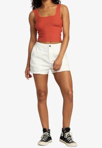 Kobieta w bezrękawnikowej, rdzawoczerwonej bluzce typu crop top, białych wysokich szortach i czarnych sneakersach za kostkę, stojąca na jednolitym tle.