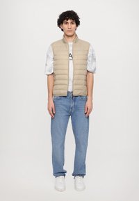 Beige puffer vest med vertikal søm, kombinert med en lys marmor-mønstret t-skjorte og blå jeans. Modellen har på seg hvite sneakers.
