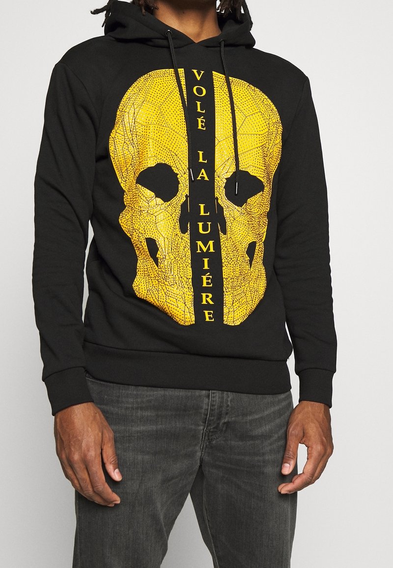 Sweat à capuche noir avec un grand crâne géométrique jaune et texte français vertical "VOLÉ LA LUMIÈRE" sur le devant porté par une personne.