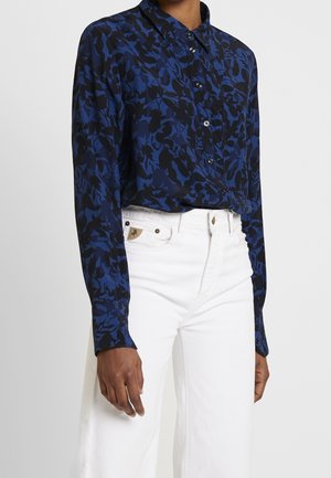 Personne portant une chemise boutonnée à manches longues bleu marine et noire à motifs floraux, rentrée dans un pantalon blanc taille haute avec des poches avant visibles.