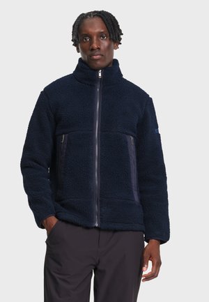 Homme portant une veste polaire bleu marine zippée, les mains dans les poches, sur un fond clair uni.