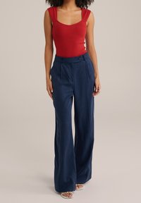 Czerwona bezrękawnikowa bluzka z kwadratowym dekoltem, połączona z spodniami o szerokich nogawkach i wysokim stanem w kolorze granatowym, z detalem guzikowym. Gładka tekstura materiału.