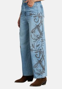 Pantalones vaqueros de pierna ancha en azul claro con bordados florales en hilo oscuro a lo largo de las costuras exteriores. Presentan una cintura alta y un cierre de cremallera.