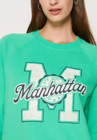 Mujer vistiendo un sudadera verde con una gran "M" blanca y el texto "Manhattan" en negro en cursiva a lo largo del pecho, con aretes de aro plateados visibles.