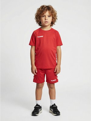 Bambino piccolo con capelli ricci in piedi, indossa maglietta e pantaloncini sportivi rossi hummel, calzini bianchi e scarpe da ginnastica nere su sfondo semplice.