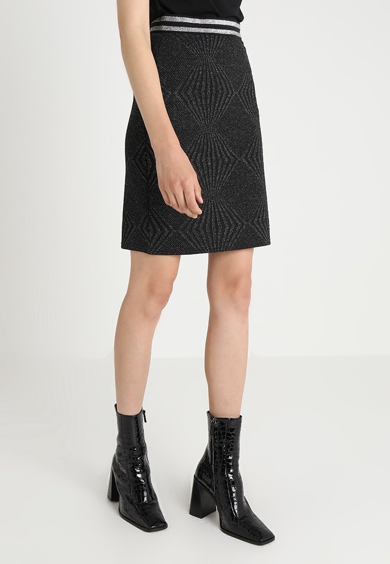 Femme portant une jupe noire texturée avec des bandes argentées à la taille et des bottines noires brillantes à talons carrés sur fond blanc.