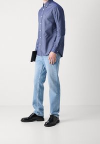 Tummansininen kauluspaita, vaaleansiniset suoralahkeiset farkut ja mustat nauhakenkä. Pitelee mustaa tablettia.