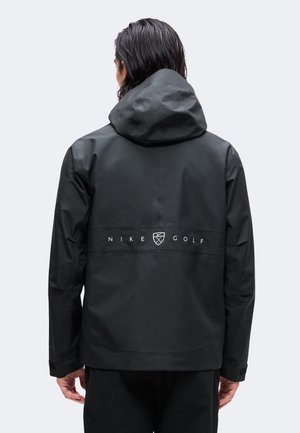 Person seen from the back wearing a black Nike Golf hooded jacket with logo and text centered on the upper back.