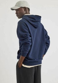 Felpa blu navy con cappuccio regolabile, polsini e orlo a coste. Indossata sopra una maglietta chiara, abbinata a pantaloni scuri. Cappellino grigio chiaro.