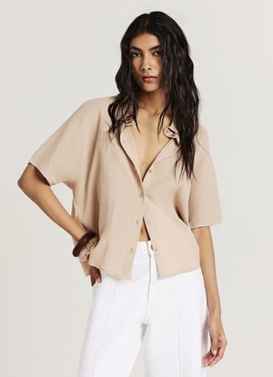 Camicia beige a maniche corte con bottoni, tessuto strutturato, colletto aperto e vestibilità ampia, abbinata a pantaloni bianchi a vita alta.