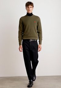 Olive green sweater over a black turtleneck, paired with black pants and black shoes. Red logo detail on the chest. Smooth texture.