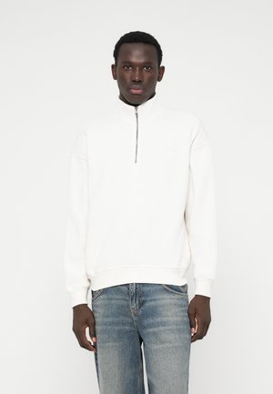 Sweat-shirt blanc zippé avec un col haut, texture douce et poignets côtelés. Associé à un jean en denim bleu clair.