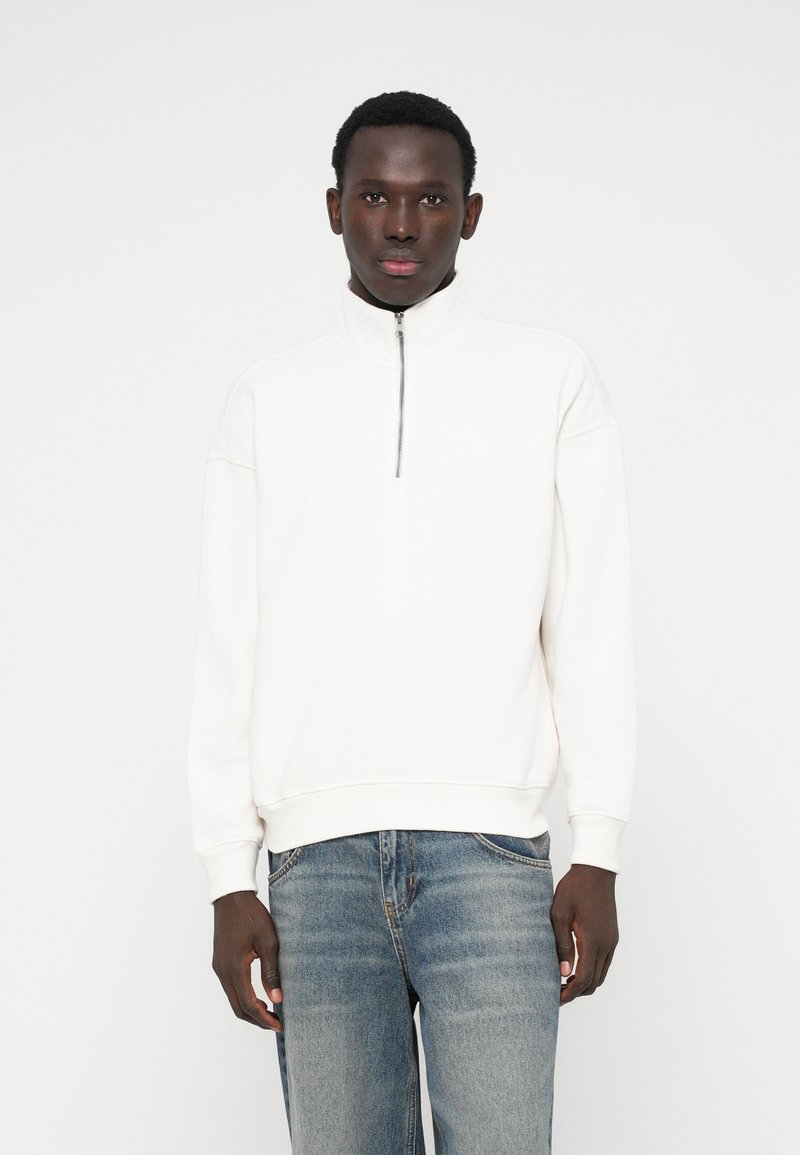 Balta svītrojuma krekls ar augstu apkakli, mīksta tekstūra un ribotām aprocēm. Apvienots ar gaiši ziliem džinsiem.