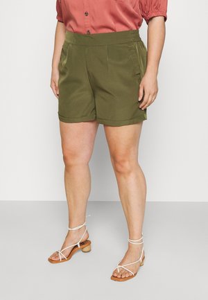 Femme portant un short sur mesure vert olive, une chemise boutonnée couleur rouille et des sandales blanches à talons avec lanières, debout sur un sol uni.
