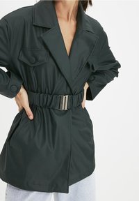 Dark green belted jacket made of smooth material, featuring large lapels, two chest pockets, and a cinched waist with a metallic buckle.
