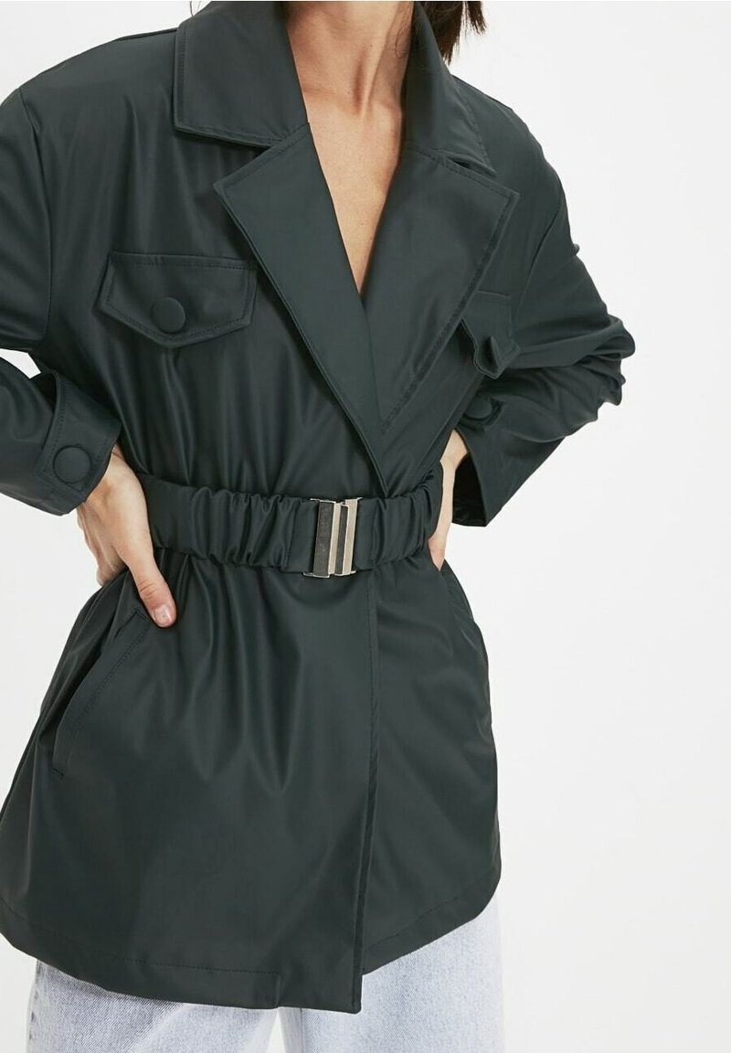 Dark green belted jacket made of smooth material, featuring large lapels, two chest pockets, and a cinched waist with a metallic buckle.