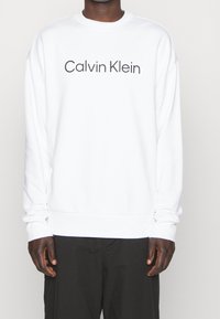 Witte sweatshirt met het "Calvin Klein" logo in een vet, donker lettertype. Ribgebreide manchetten en zoom met een losse pasvorm. Zachte stoftextuur.