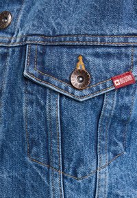 Denimska tkanina u plavoj boji s bakrenim dugmadima, sadrži džep na prsima i narančaste šavove. Vidljiva je crvena oznaka s natpisom "BIG STAR".