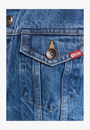 Tissu en denim bleu avec des boutons en cuivre, comportant une poche poitrine et des coutures oranges. Une étiquette rouge portant "BIG STAR" est visible.