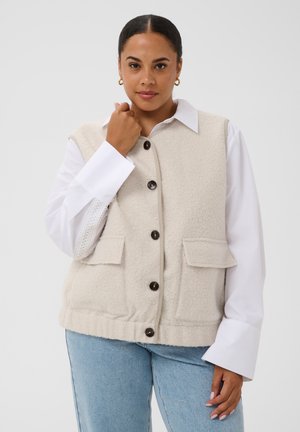 Femme portant un gilet en laine beige boutonné sur une chemise blanche à larges poignets, associé à un jean bleu clair, debout devant un fond uni.