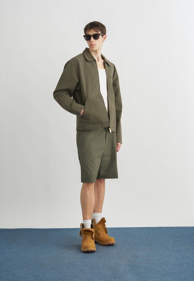 Giovane uomo con giacca verde scuro e pantaloncini, canottiera bianca, stivali da lavoro color sabbia, occhiali da sole, in piedi con una mano in tasca su un pavimento blu.