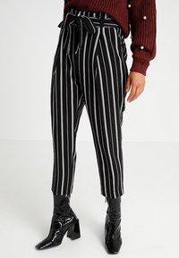Černobílé pruhované kalhoty s vysokým páskem, vázacím opaskem a zúženými nohavicemi, doplněné lesklými černými kotníkovými botami.