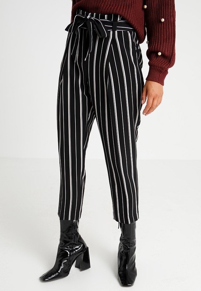 Černobílé pruhované kalhoty s vysokým páskem, vázacím opaskem a zúženými nohavicemi, doplněné lesklými černými kotníkovými botami.