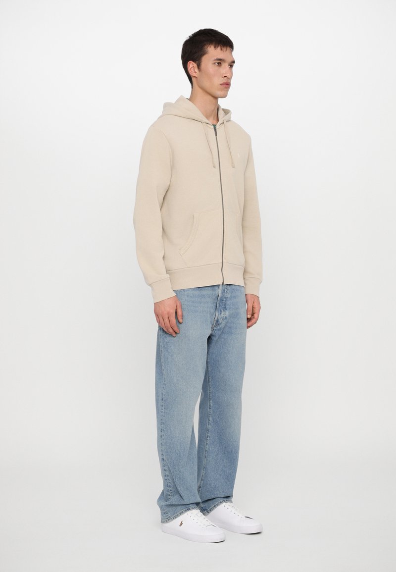 Beige zip-up hoodie van zachte stof, met een voorzak, een capuchon met trekkoord en geribbelde manchetten, gecombineerd met lichtblauwe jeans en witte sneakers.