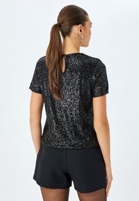 Zwarte pailletten top met korte mouwen, voorzien van een keyhole-detail aan de achterkant en een gladde textuur, gecombineerd met zwarte shorts.