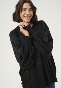 Černá, oversized blůza s dlouhými nabíranými rukávy a smockovanými manžetami. Hladká textura a knoflíková léga vpředu. Casual střih.