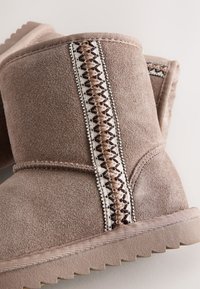 Stivali in camoscio taupe invernali con dettaglio cuciture a motivo zigzag e suole in gomma scanalate, mostrati in primo piano su uno sfondo neutro.