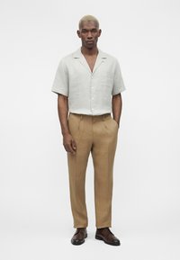 Άνδρας με ξανθά κοντά μαλλιά, που φοράει ανοιχτό γκρι κοντομάνικο πουκάμισο με κουμπιά, μπεζ παντελόνι και σκούρα καφέ επίσημα παπούτσια, στέκεται όρθιος.