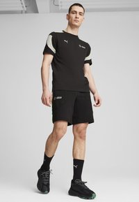 Musta urheilupaita vihreillä ja harmailla yksityiskohdilla, paritettuna mustien shortsejen kanssa, joissa on teksturoitu kuvio ja logo. Mustat kengät ja sukat viimeistelevät kokonaisuuden.