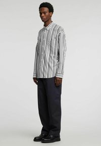 Długorękawowa koszula w czarno-białe paski, z zapięciem na guziki, kieszenią na piersi oraz kontrastującymi wzorami pasków. Noszona z ciemnymi spodniami.
