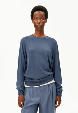 Femme aux cheveux courts et bouclés portant un pull bleu à manches longues et un pantalon bleu ample, debout sur un fond blanc.