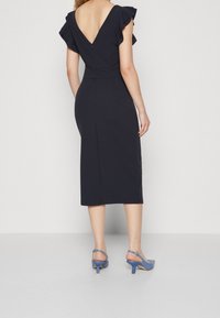 Mekko, jossa on laivastonsininen väri, V-mallinen selkä, leijaillat ja vartalonmyötäinen siluetti. Kangas on sileä, polvien pituinen, ja asusteena ovat vaaleansiniset korkokengät.