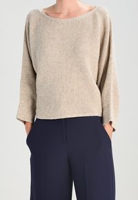 Suéter largo de manga larga en beige con un escote amplio, combinado con pantalones azul marino de corte holgado, con las manos en los bolsillos.